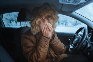 man in winter coat blowing into his hand in a broken down car during a snow storm