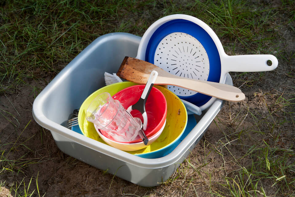 dirty dishes piled in a plastic tub sitting on dry grass