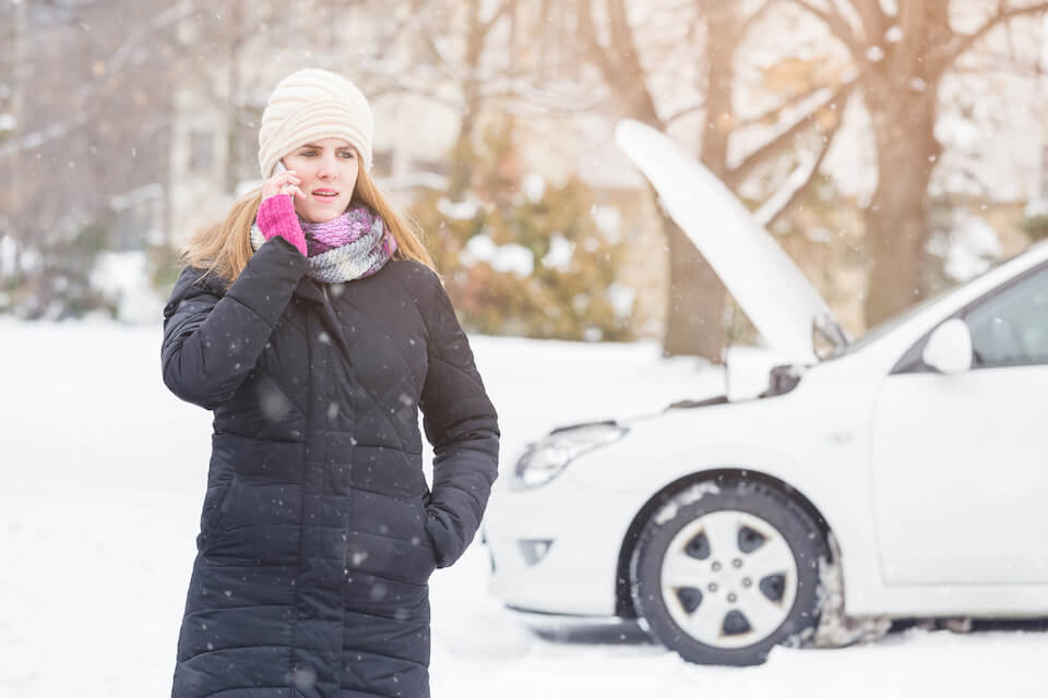 woman in a heavy coat and hat on the phone as she stands in front of her broken down car with the hood open in a snow storm