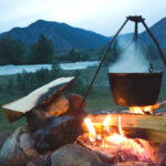 metal pot hanging on a tripod over a fire outdoors in front of a lake and mountain backdrop