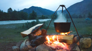 metal pot hanging on a tripod over a fire outdoors in front of a lake and mountain backdrop