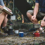 two people sitting in camping chairs cooking in a small pan over a portable stove in the outdoors